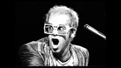 Elton John playing piano with receding hair and big specs